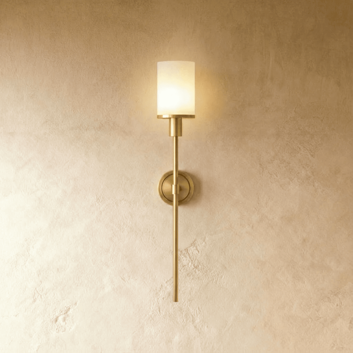 Clérachic brass wall sconce with a glowing white cylinder shade, round backplate and slim rod on a limewash wall.