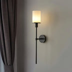 Clérachic black wall sconce with a warm glowing cylinder shade on a grey wall beside a taupe curtain.