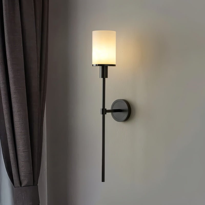 Clérachic black wall sconce with a warm glowing cylinder shade on a grey wall beside a taupe curtain.