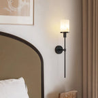 Clérachic black wall sconce with a veined white stone-look cylinder shade, lit beside an arched bedhead and linen curtain.