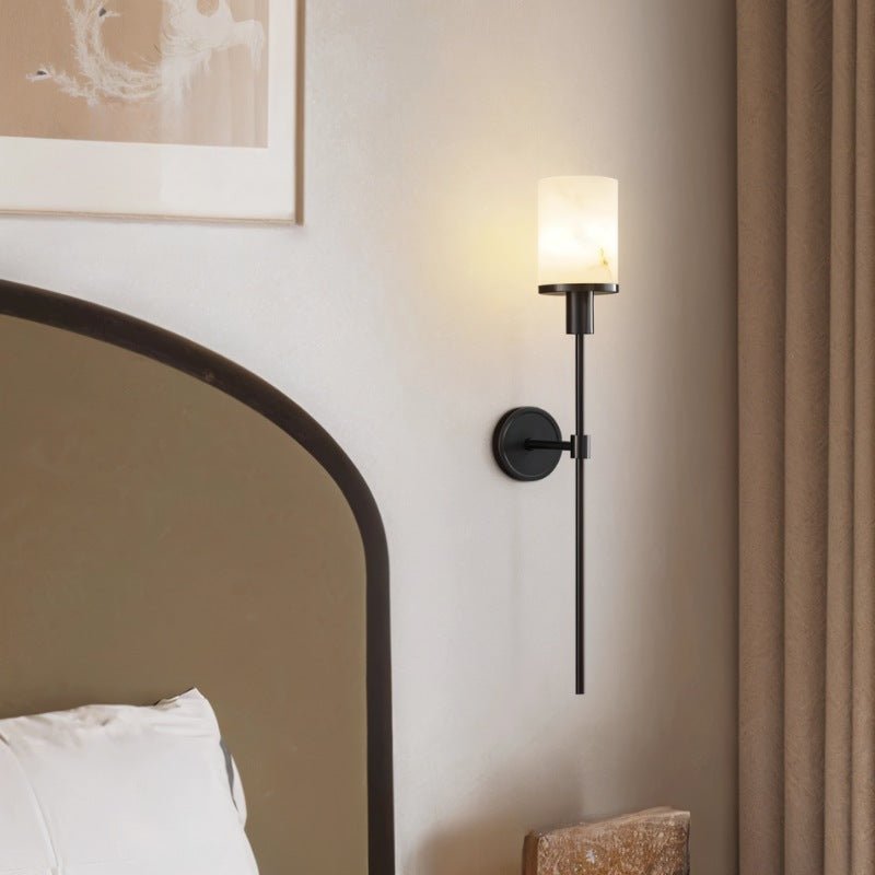 Clérachic black wall sconce with a veined white stone-look cylinder shade, lit beside an arched bedhead and linen curtain.