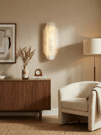A lit Clérachic alabaster wall sconce on a cream wall above a walnut sideboard, with dried grasses, a pale bouclé armchair and a floor lamp beside it.