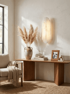 Clérachic alabaster sconce glowing on a pale wall above a wooden console table, with pampas grasses in a stone vase and a woven throw on a chair.