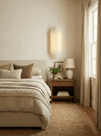 A Clérachic wall sconce lit beside a bedroom window with linen curtains, above a bed with neutral bedding and a wooden bedside table.