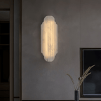 A Clérachic alabaster sconce glowing on a dark grey plaster wall, with a framed screen and a slim vase of dried stems below.