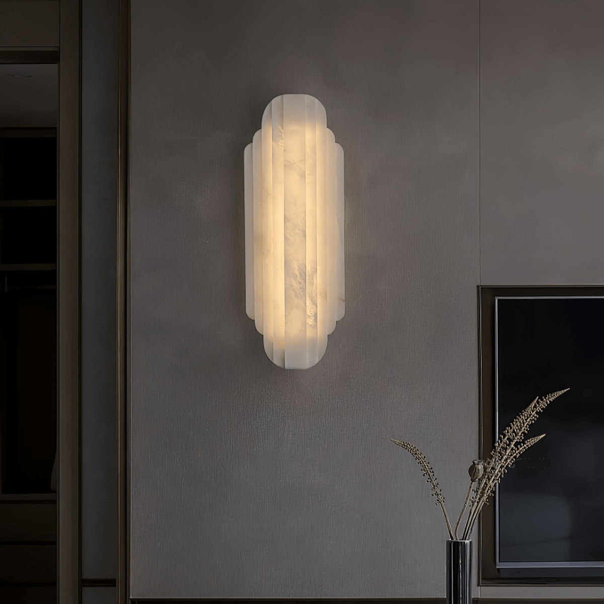 A Clérachic alabaster sconce glowing on a dark grey plaster wall, with a framed screen and a slim vase of dried stems below.