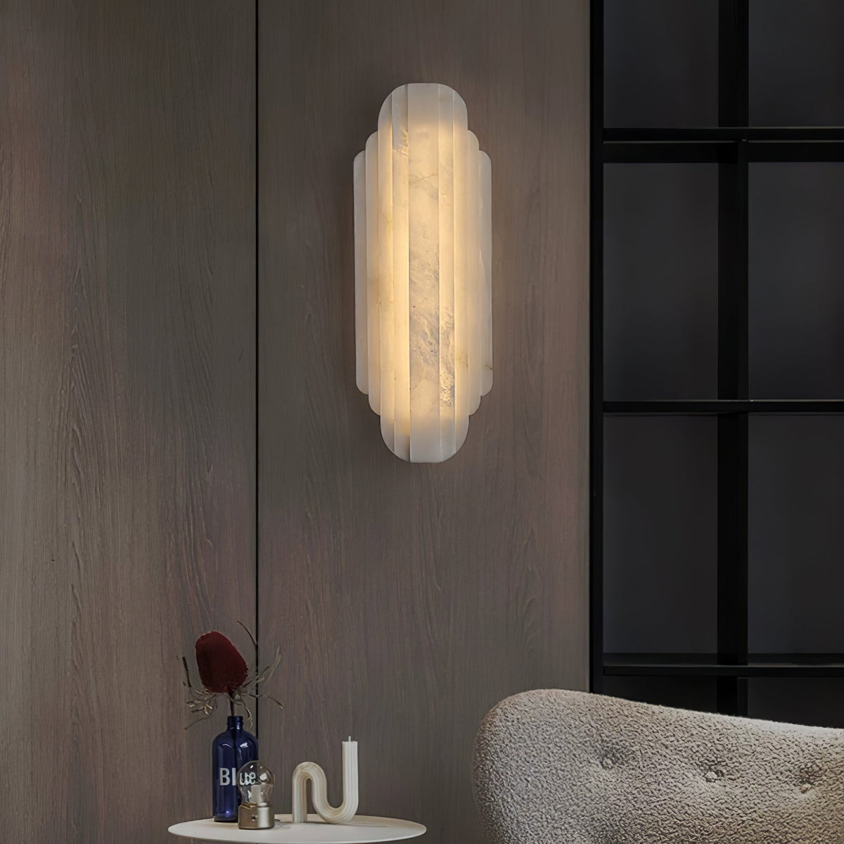 Clérachic alabaster sconce lit on a dark wood-panelled wall beside a black-framed window, above a small table and a bouclé chair.