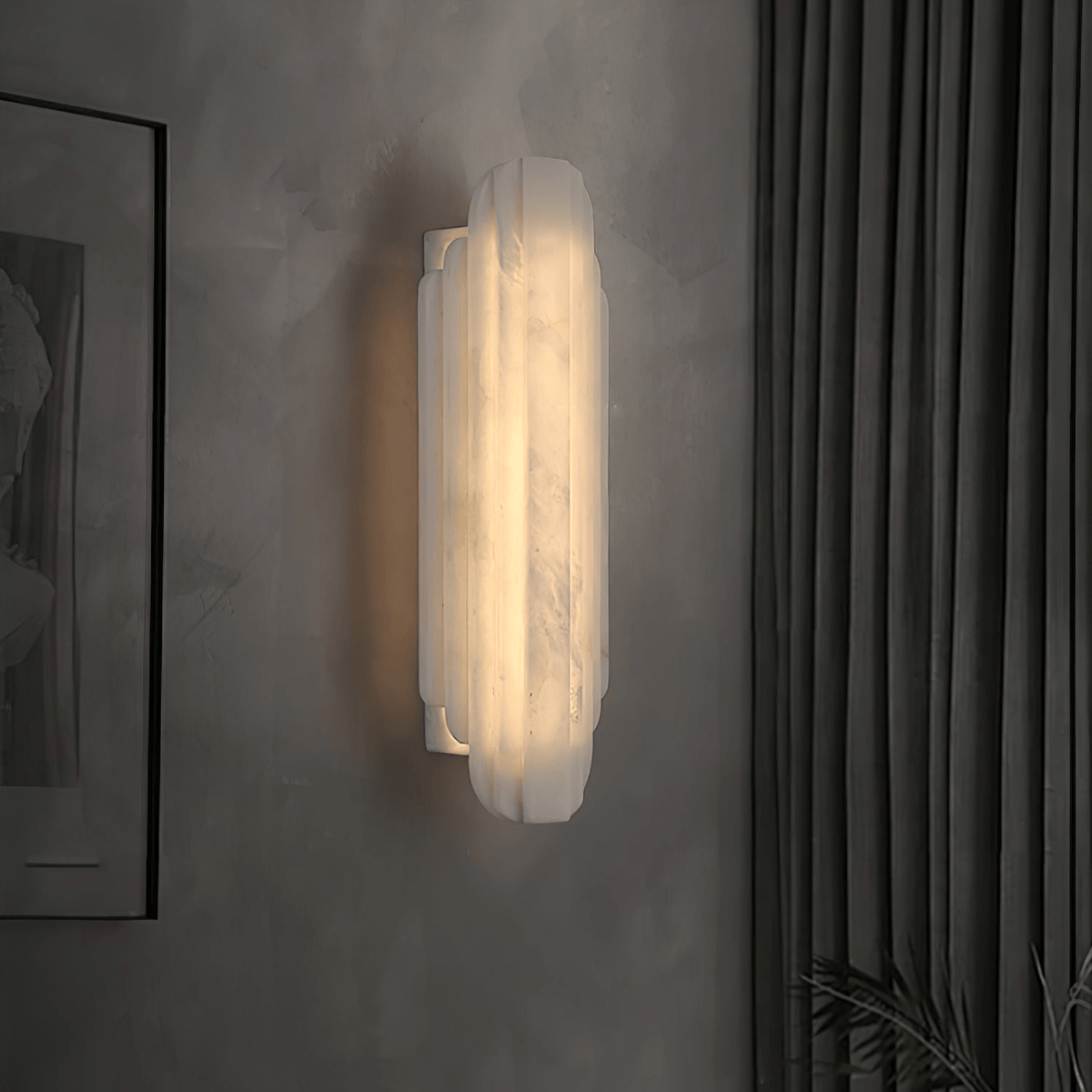 Side-angled view of this Clérachic alabaster sconce lit on a grey wall beside a dark curtain, showing its stepped edge and slim wall plate.