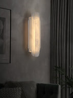 A tall, lit Clérachic alabaster wall sconce on a grey wall between a framed picture and a dark curtain, above a tufted armchair.