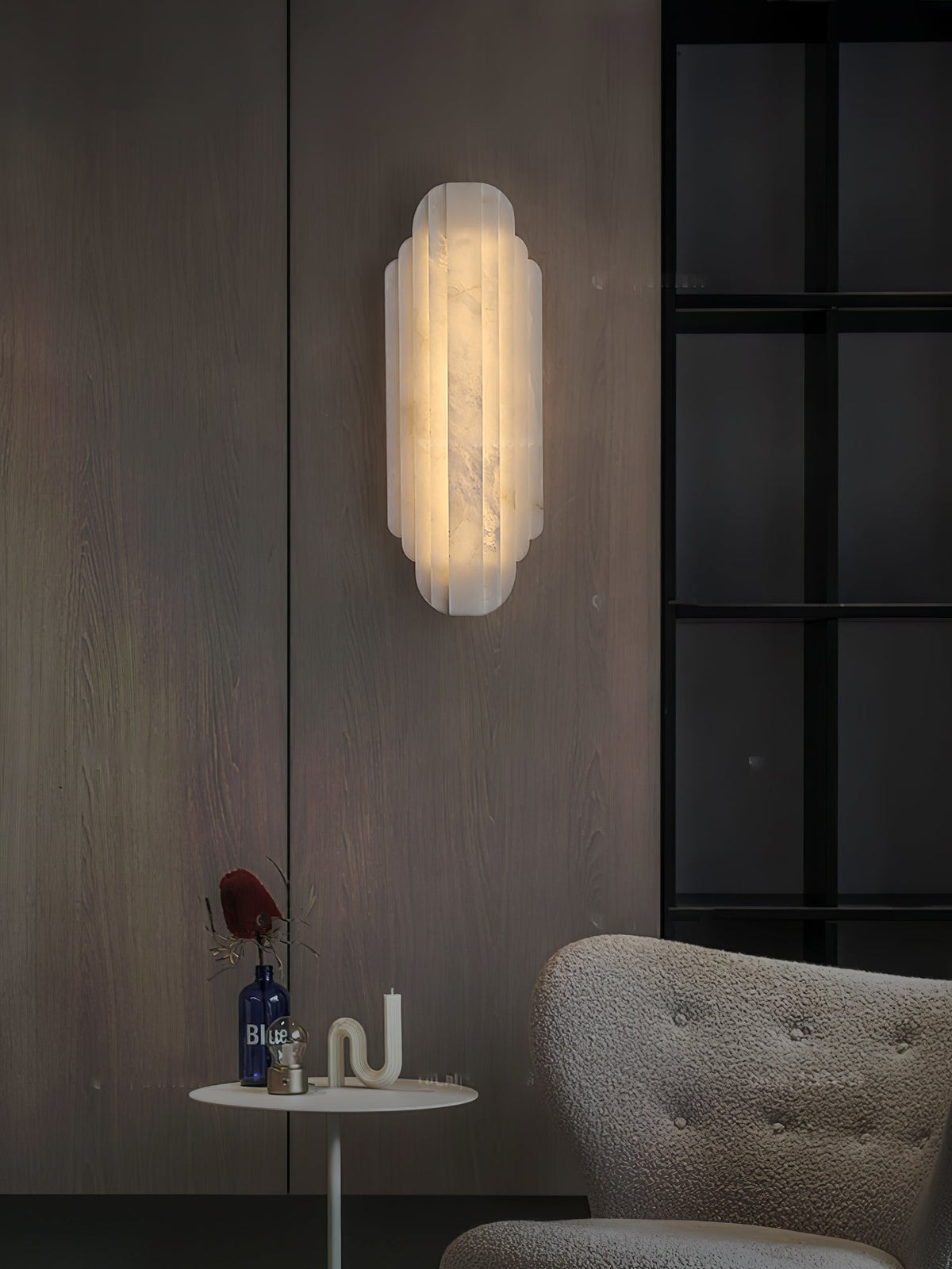 Clérachic alabaster sconce glowing on a wood-panelled wall, with a black-framed window and a small round table below.