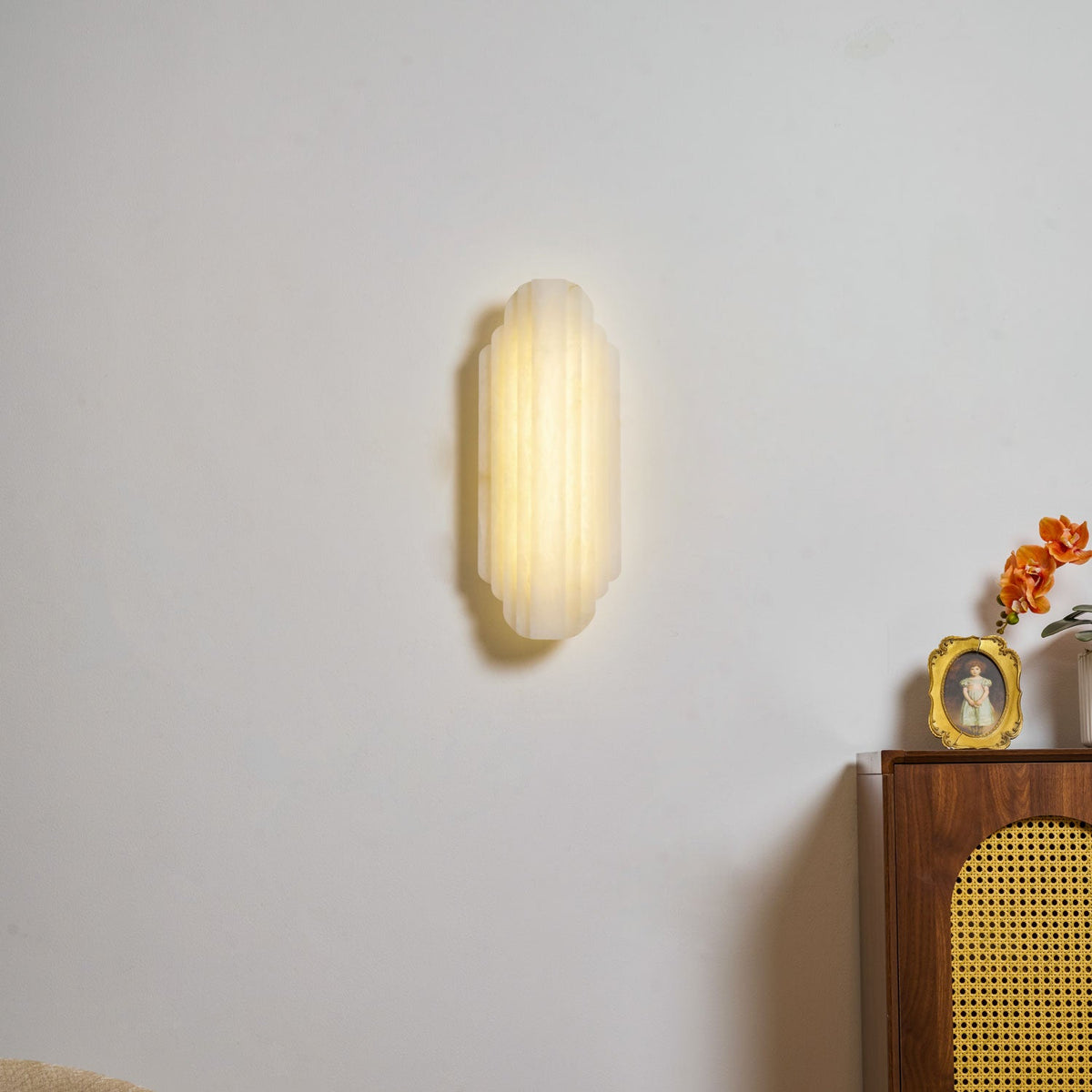 The lit Clérachic sconce on a pale wall, its ribbed alabaster glowing warm above a cane-fronted walnut cabinet with a gold frame.