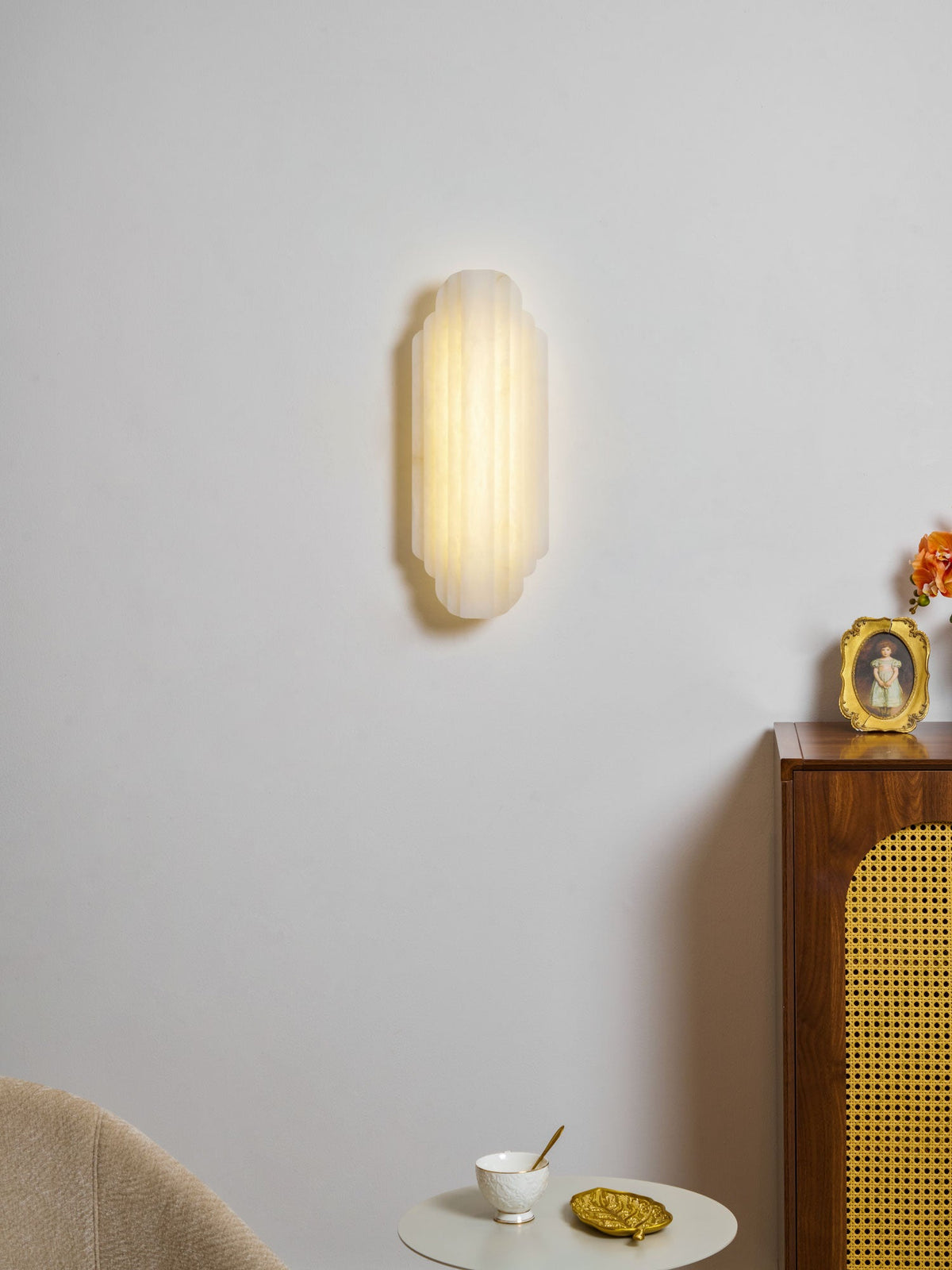 A lit Clérachic wall sconce on a pale grey wall, above a cane-fronted cabinet and a small white table with a cup.