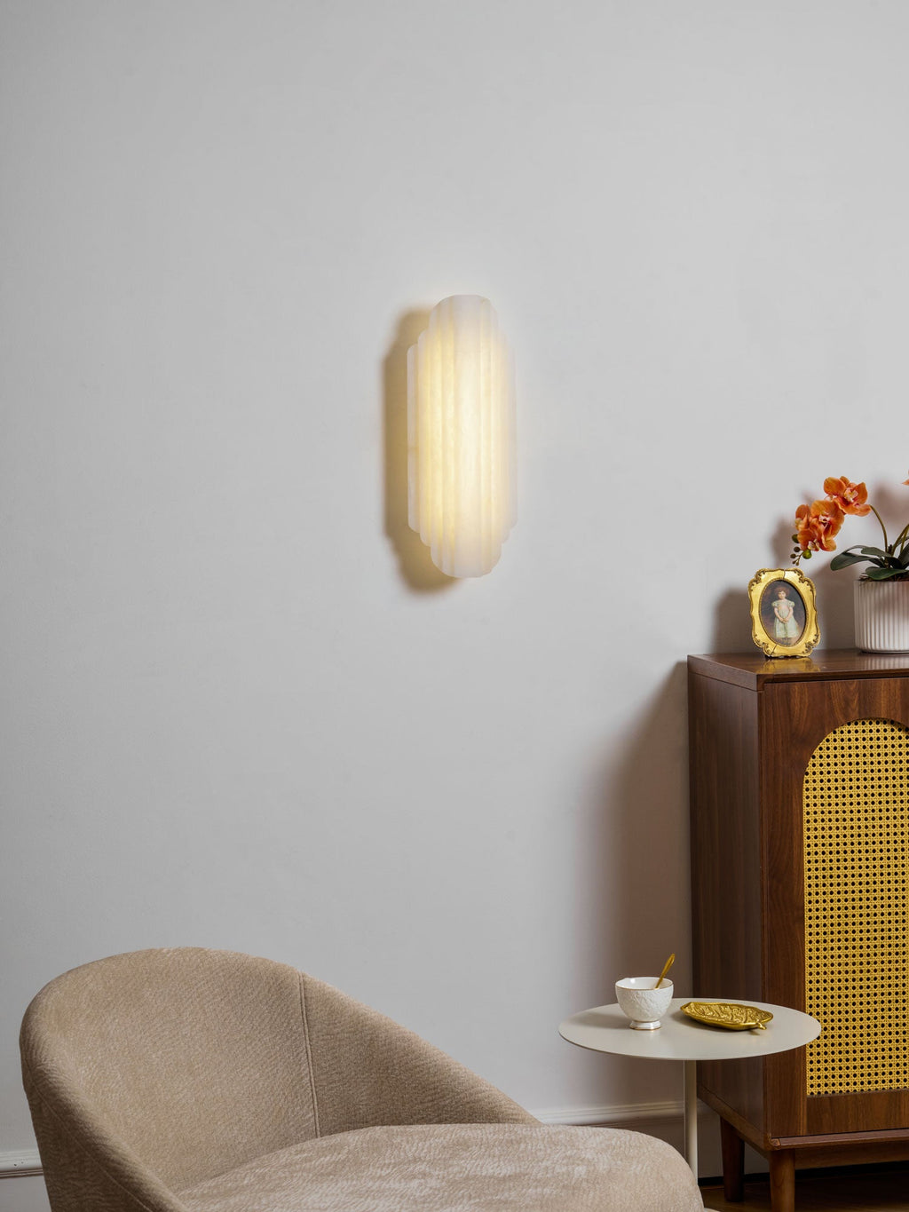A lit Clérachic sconce hangs high on a pale wall above a cane-fronted cabinet, with a beige armchair and a round side table below.