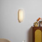 A closer view of the lit Clérachic alabaster sconce on a pale wall, with a cane-fronted cabinet and orange flowers to the right.