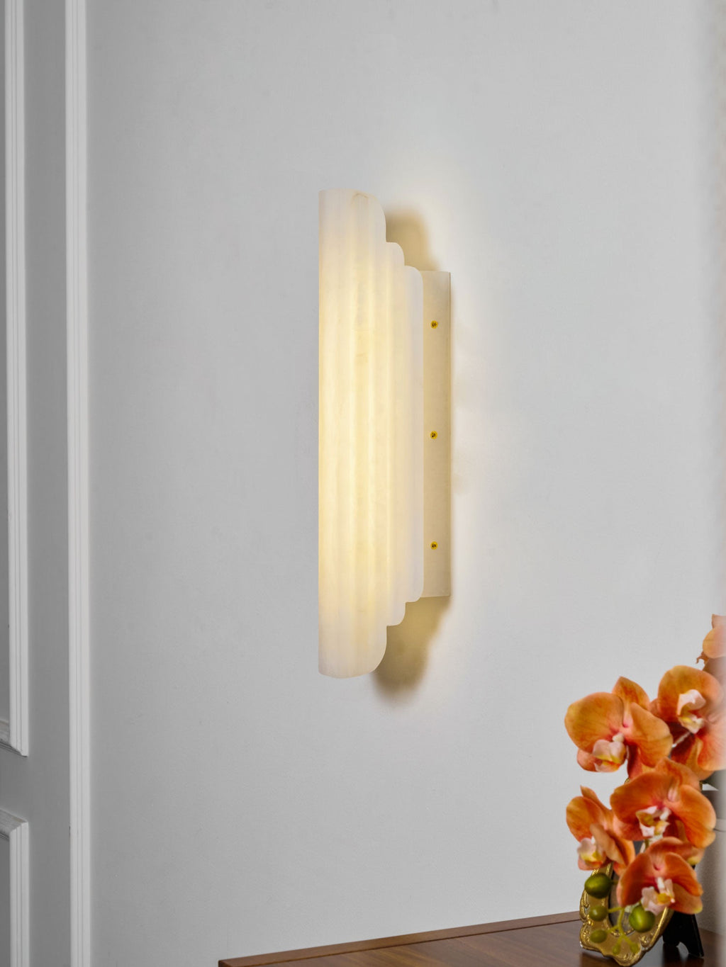 Side view of this Clérachic alabaster sconce lit on a white wall, showing the stepped depth of its ribbed shade and its slim brass-dotted back plate.