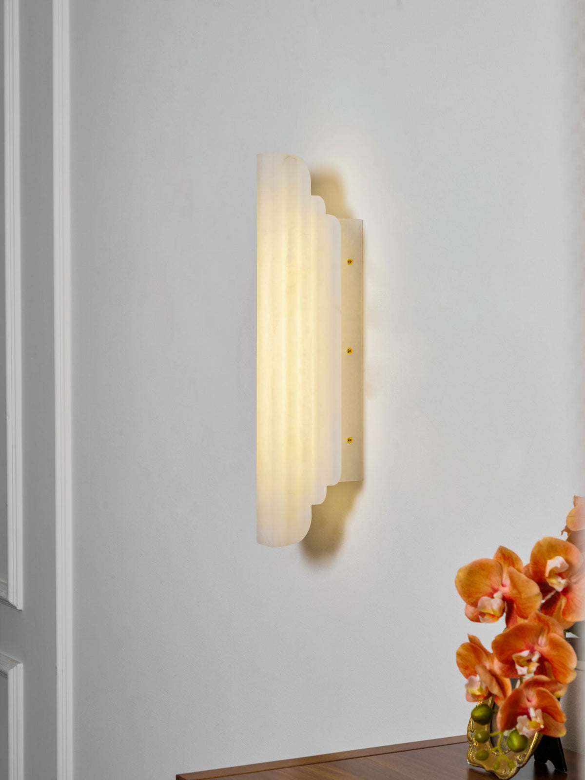 Side view of this Clérachic alabaster sconce lit on a white wall, showing the stepped depth of its ribbed shade and its slim brass-dotted back plate.