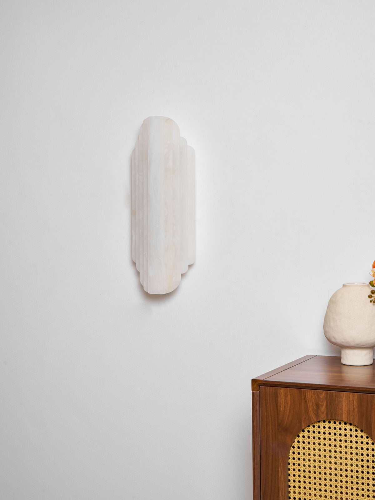An unlit Clérachic alabaster sconce on a white wall above a walnut cabinet with a cane front and a rough-textured cream vase.