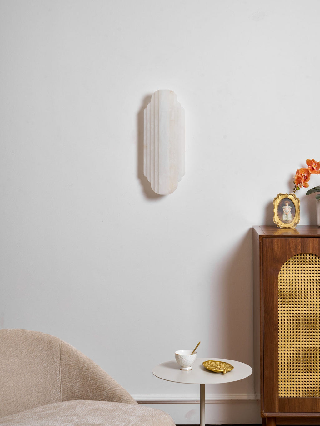 The unlit Clérachic alabaster wall sconce on a white wall, above a cane-fronted cabinet and a small round table with a cup and dish.