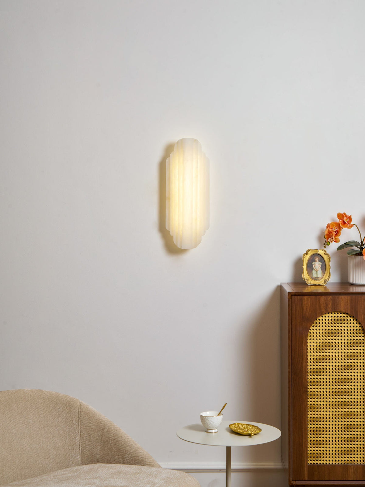 A lit Clérachic alabaster sconce on a white wall, glowing warm above a cane-fronted cabinet, a beige chair and a small round table.