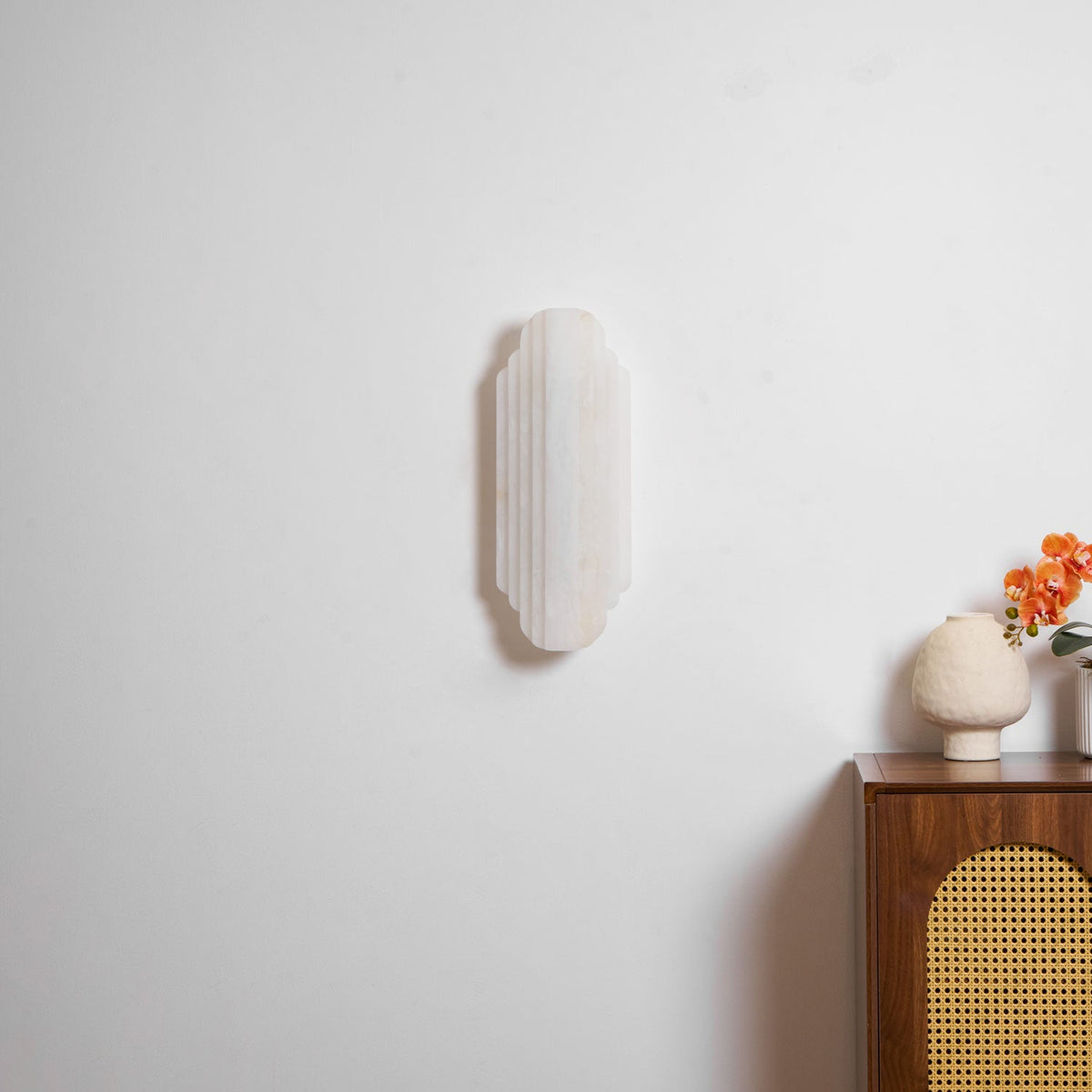 A slightly closer view of the unlit Clérachic sconce on a white wall, with a pale vase and orange flowers on the cabinet below.