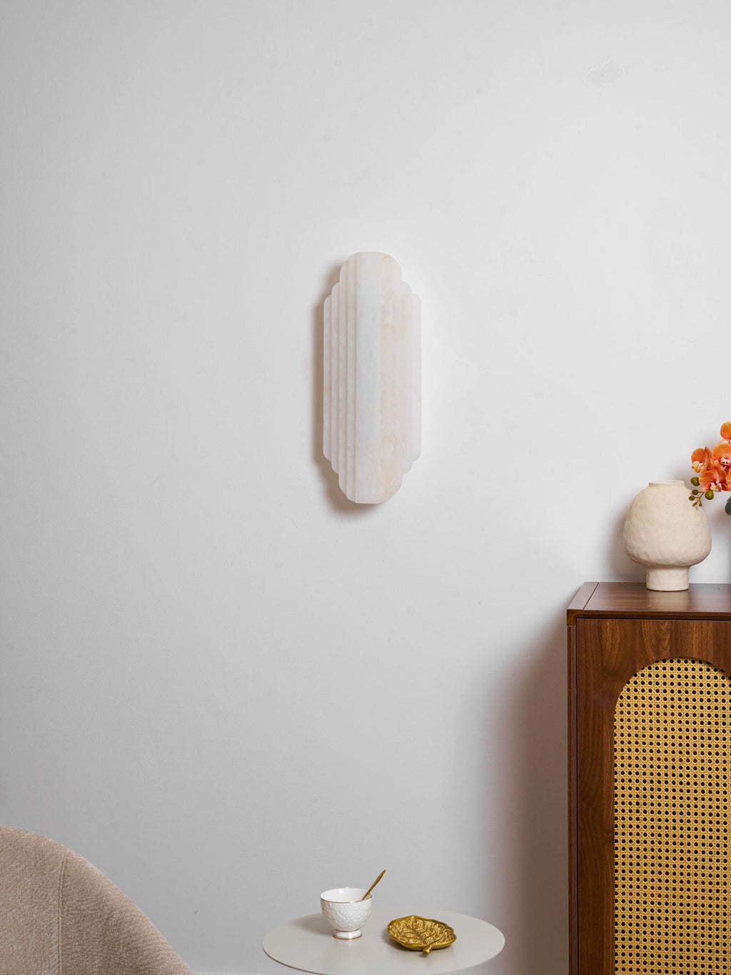 The unlit Clérachic alabaster sconce on a white wall, with a ceramic vase on a cane-fronted cabinet and a small table with a cup.