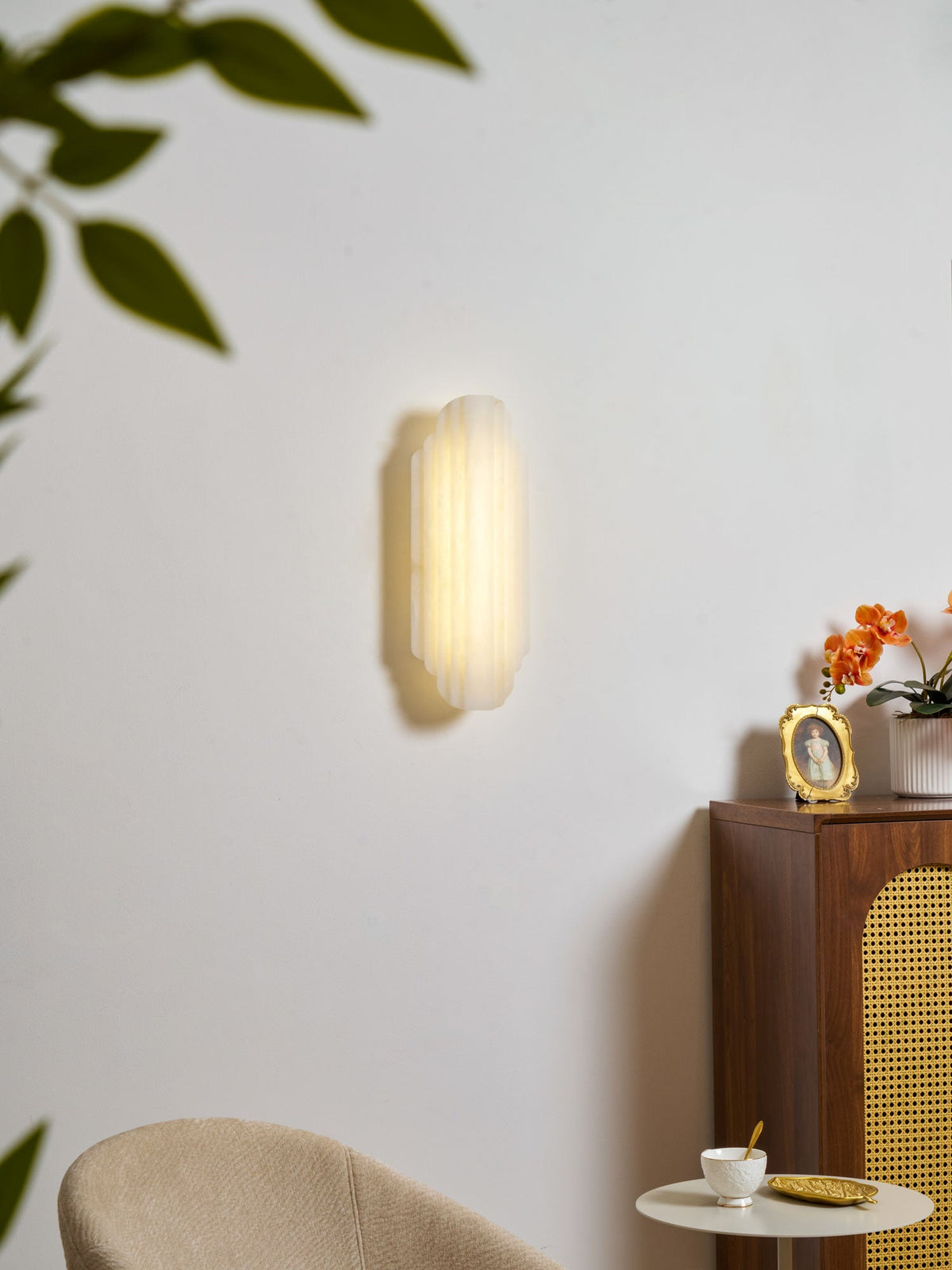 A lit Clérachic alabaster wall sconce on a white wall, with green leaves blurred at the top and a cane-fronted cabinet below.