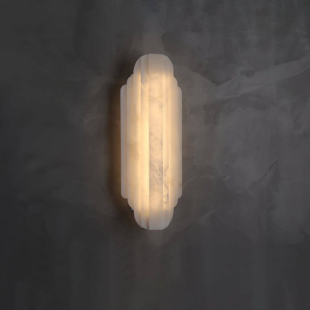 A shorter Clérachic alabaster sconce lit on a dark grey plaster wall, its ribbed shade glowing warm.