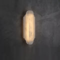 A shorter Clérachic alabaster sconce lit on a dark grey plaster wall, its ribbed shade glowing warm.