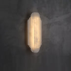 A shorter Clérachic alabaster sconce lit on a dark grey plaster wall, its ribbed shade glowing warm.