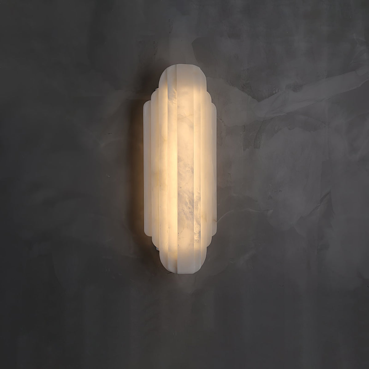 A shorter Clérachic alabaster sconce lit on a dark grey plaster wall, its ribbed shade glowing warm.