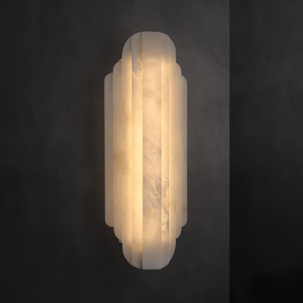 A tall Clérachic alabaster sconce lit on a dark grey wall, its stepped ribbed shade glowing warm from top to bottom.
