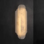 A tall Clérachic alabaster sconce lit on a dark grey wall, its stepped ribbed shade glowing warm from top to bottom.