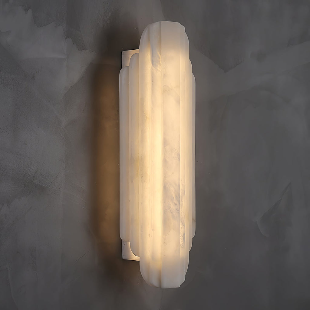 Three-quarter view of this Clérachic alabaster sconce lit on a dark grey wall, with the slim white back plate visible behind the shade.