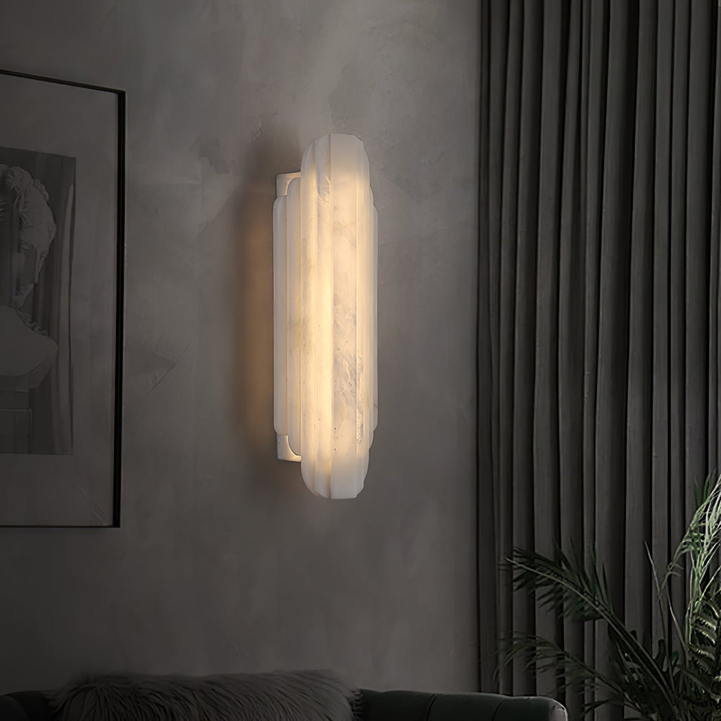 A Clérachic alabaster sconce lit on a grey wall beside a dark curtain and a plant, seen from the side.