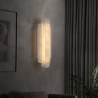 A Clérachic alabaster sconce lit on a grey wall beside a dark curtain and a plant, seen from the side.
