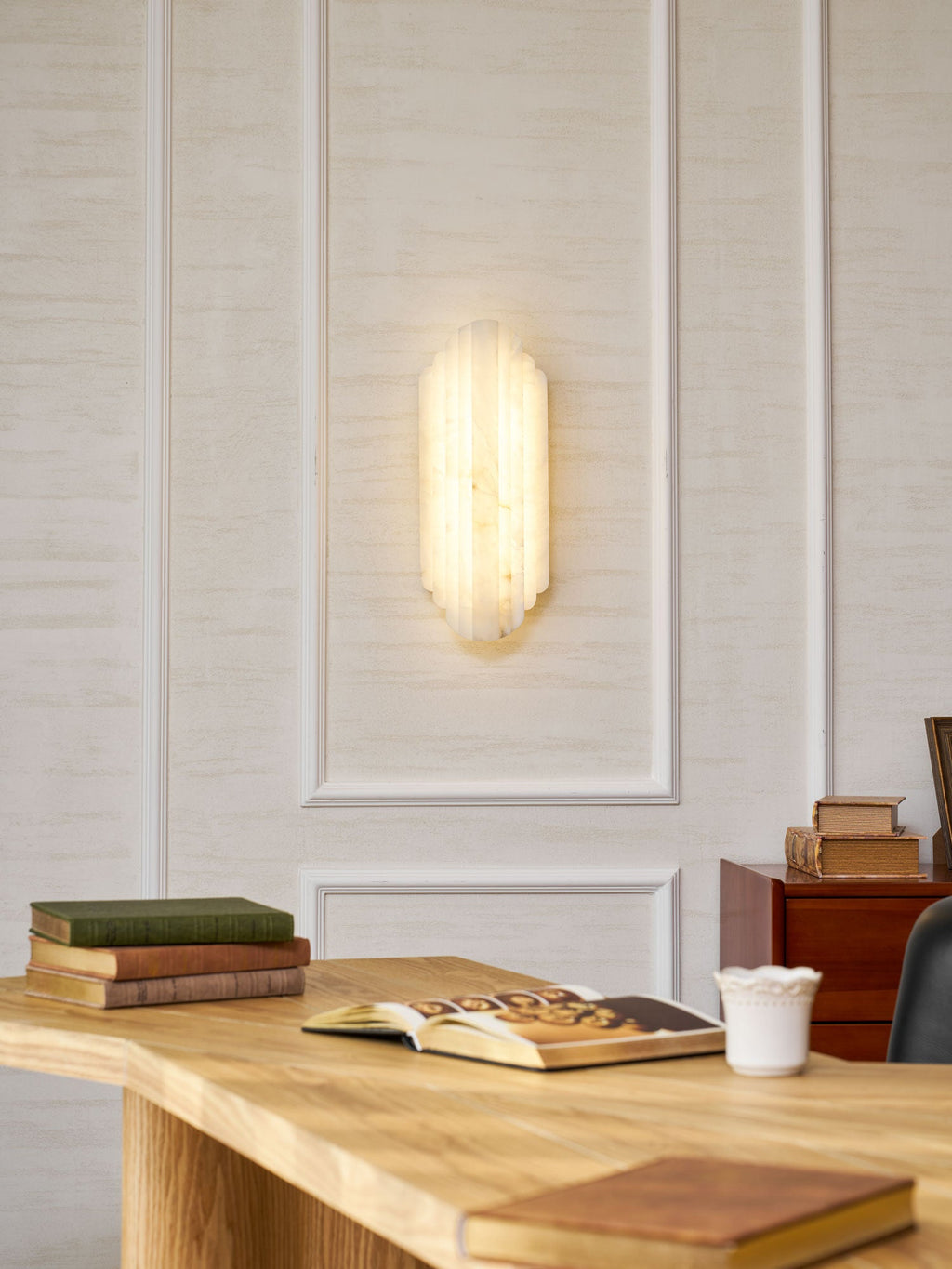 A lit Clérachic alabaster sconce on a panelled cream wall above a pale wooden desk with books and a cup.