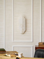 The unlit Clérachic alabaster sconce on a panelled cream wall, above a desk with books, a cup and a black leather chair.