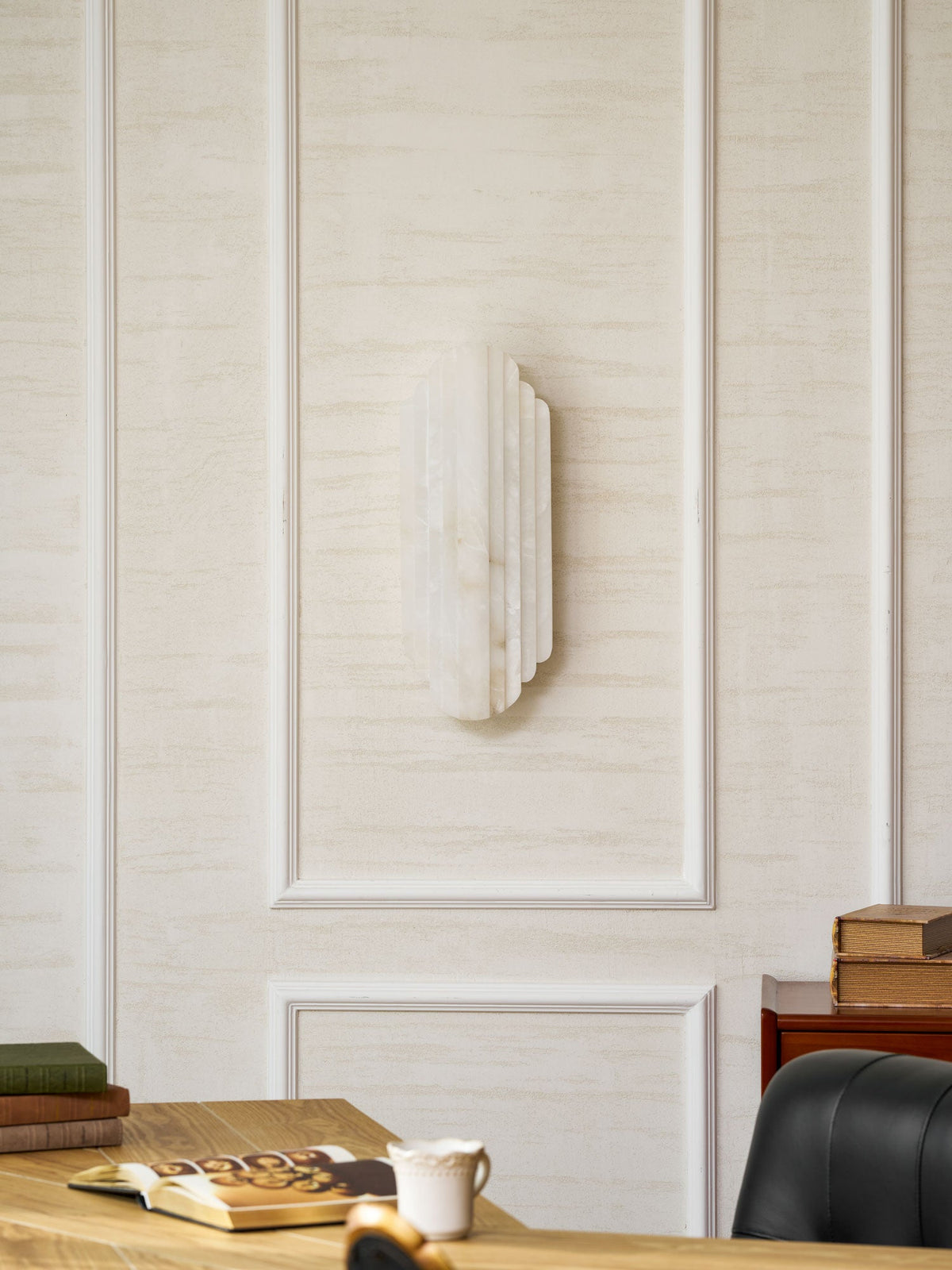The unlit Clérachic alabaster sconce on a panelled cream wall, above a desk with books, a cup and a black leather chair.