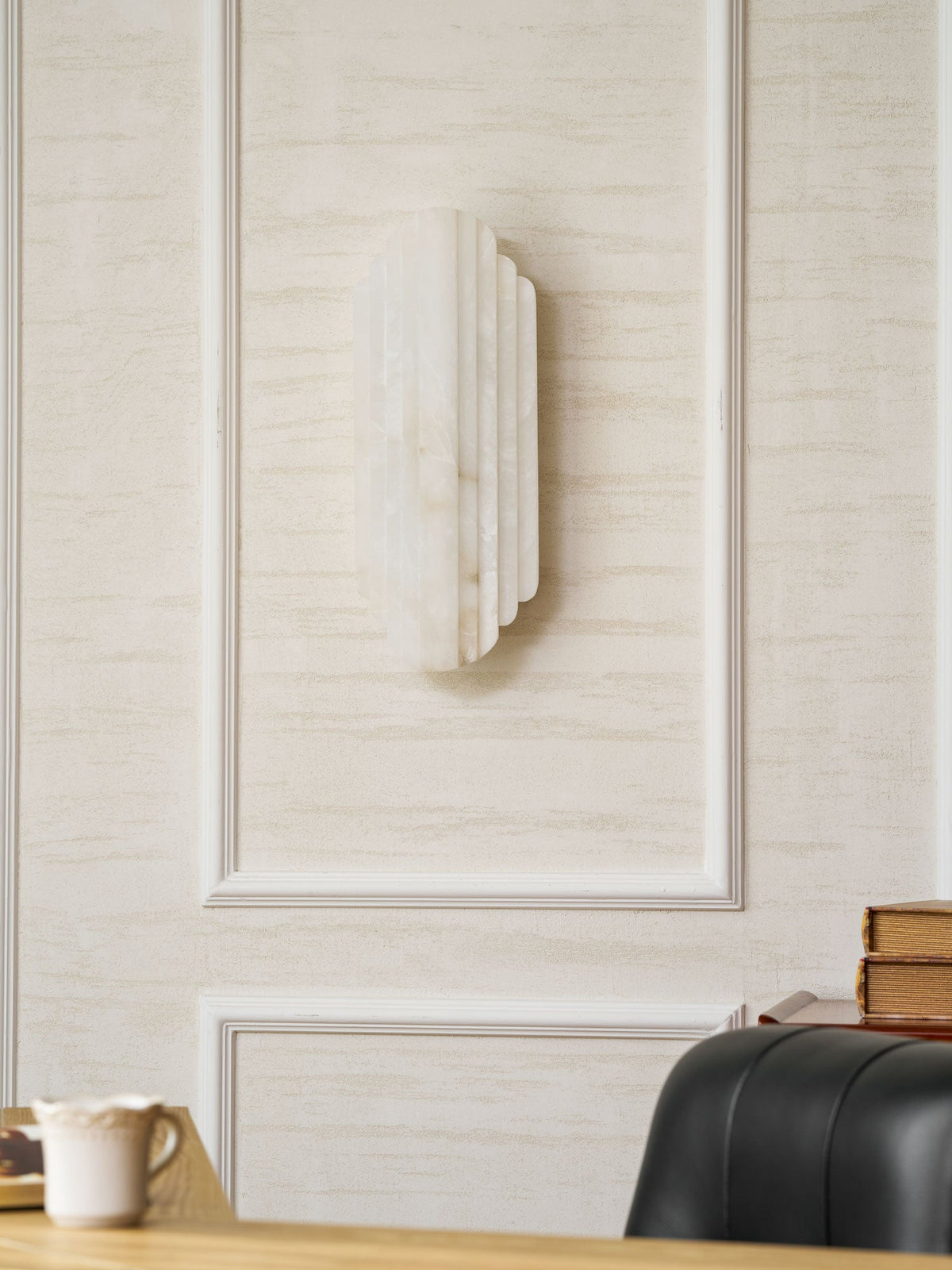 An unlit Clérachic alabaster sconce on a panelled cream wall, seen from the side above a desk and a black leather chair.