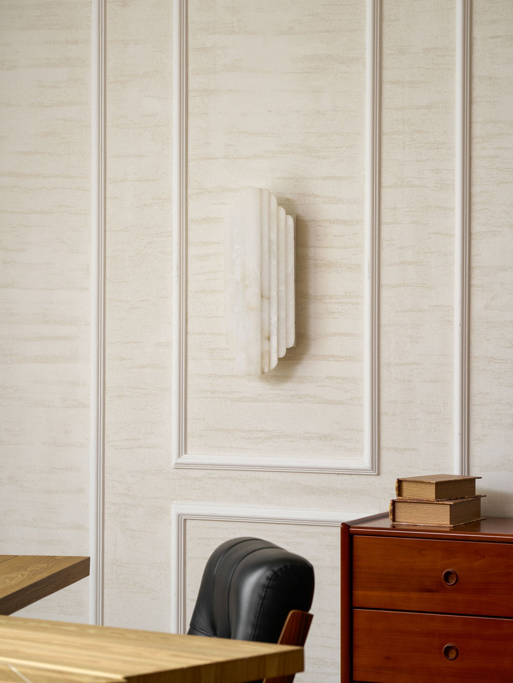 The unlit Clérachic alabaster sconce on a panelled wall, with a black leather chair and a chest of drawers below it.