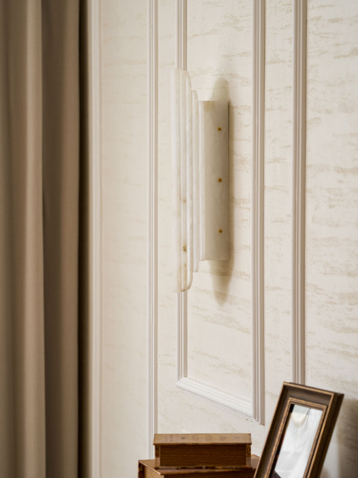 Side view of the unlit Clérachic alabaster sconce on a panelled wall beside a taupe curtain, with a framed picture below.