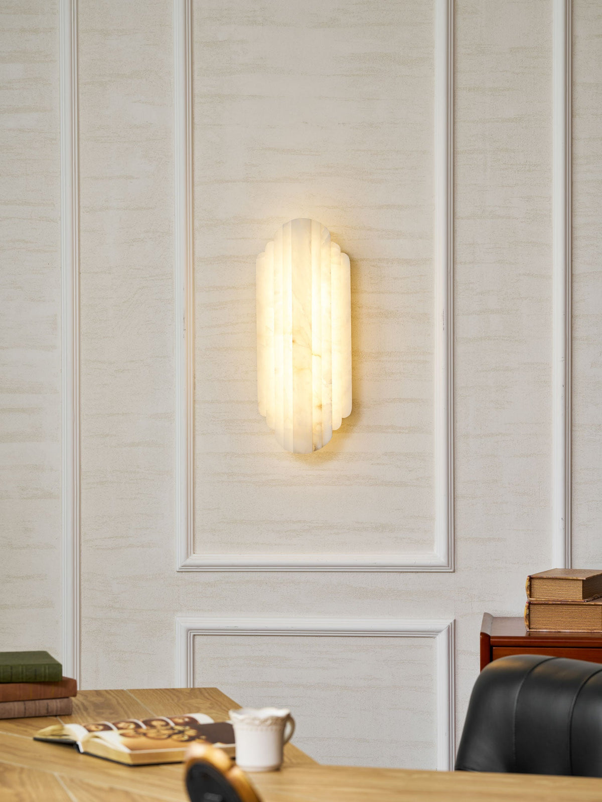 A lit Clérachic alabaster sconce glowing warm on a panelled cream wall, above a desk with an open book and a white cup.