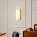 The lit Clérachic alabaster sconce on a panelled wall, glowing warm above a chest of drawers with books and a framed picture.