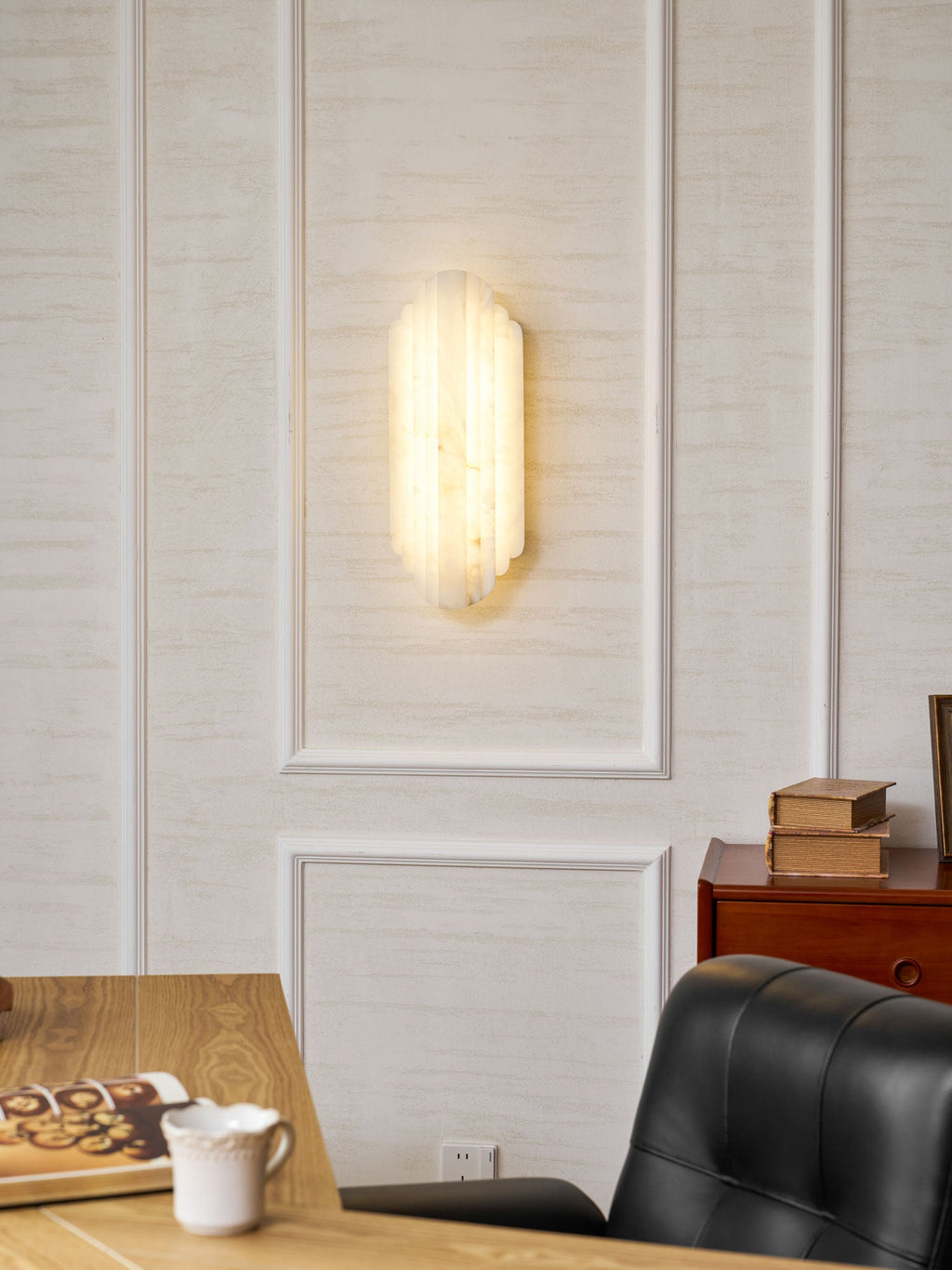 A lit Clérachic alabaster sconce on a panelled wall, with a desk, an open book, a cup and a black leather chair below.