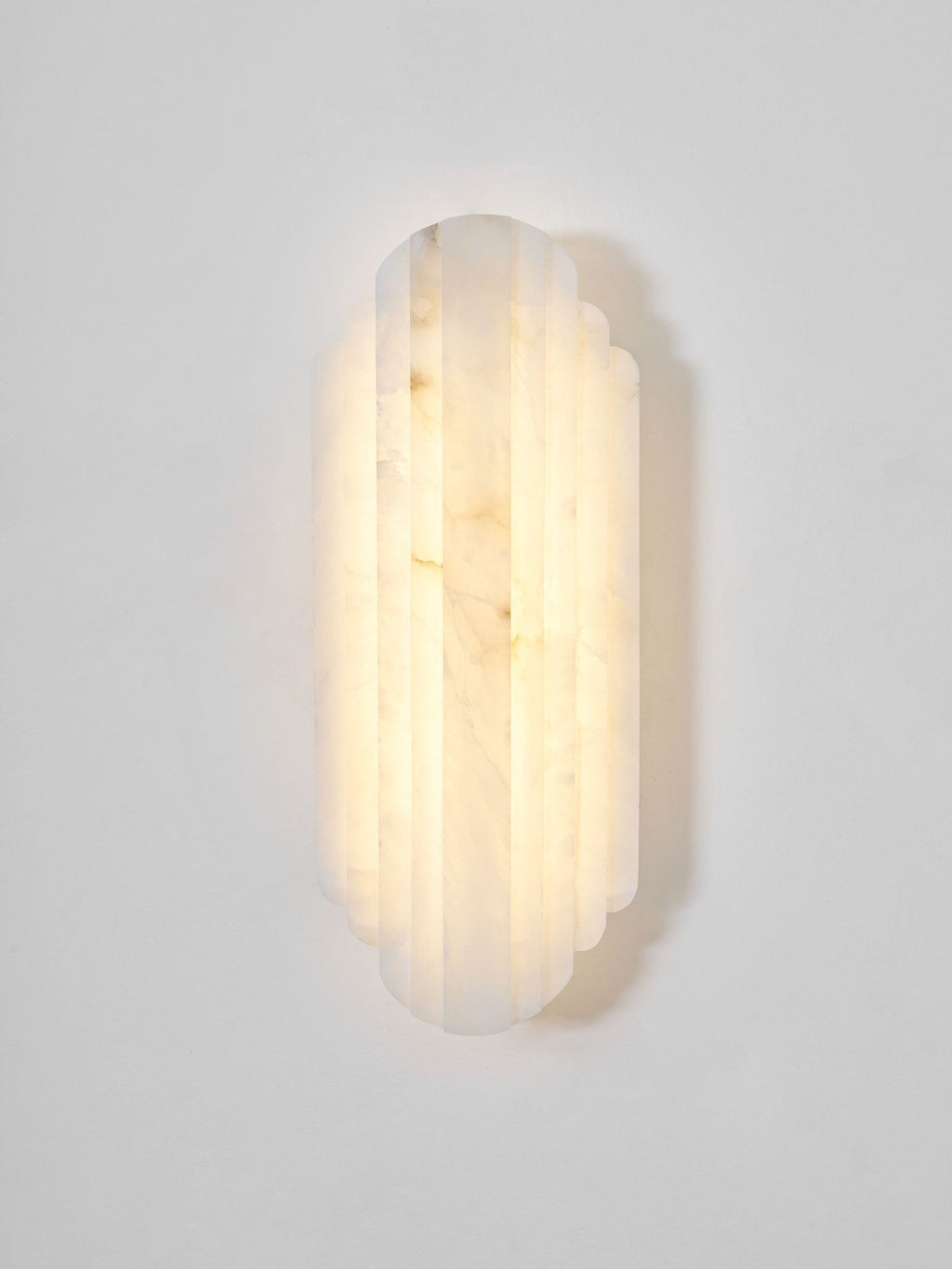 Front view of this lit Clérachic alabaster sconce on a pale grey wall, its stepped ribbed shade glowing warm with soft veining.
