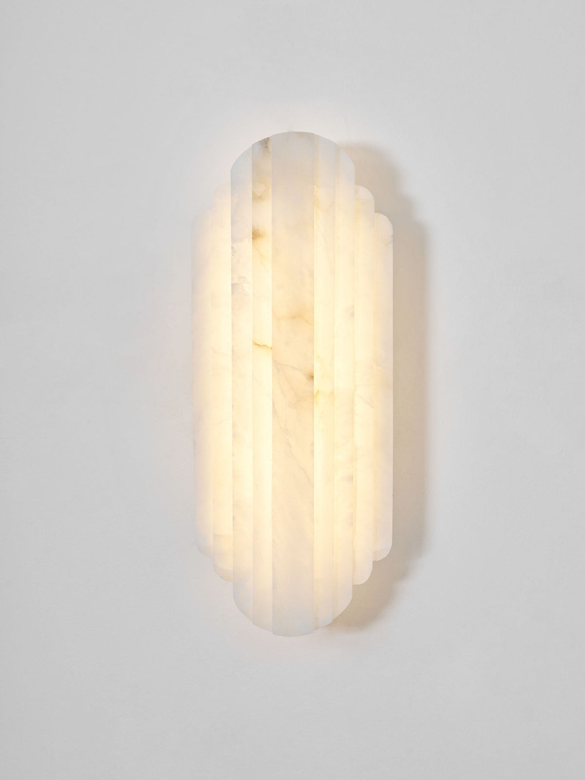 Front view of this lit Clérachic alabaster sconce on a pale grey wall, its stepped ribbed shade glowing warm with soft veining.