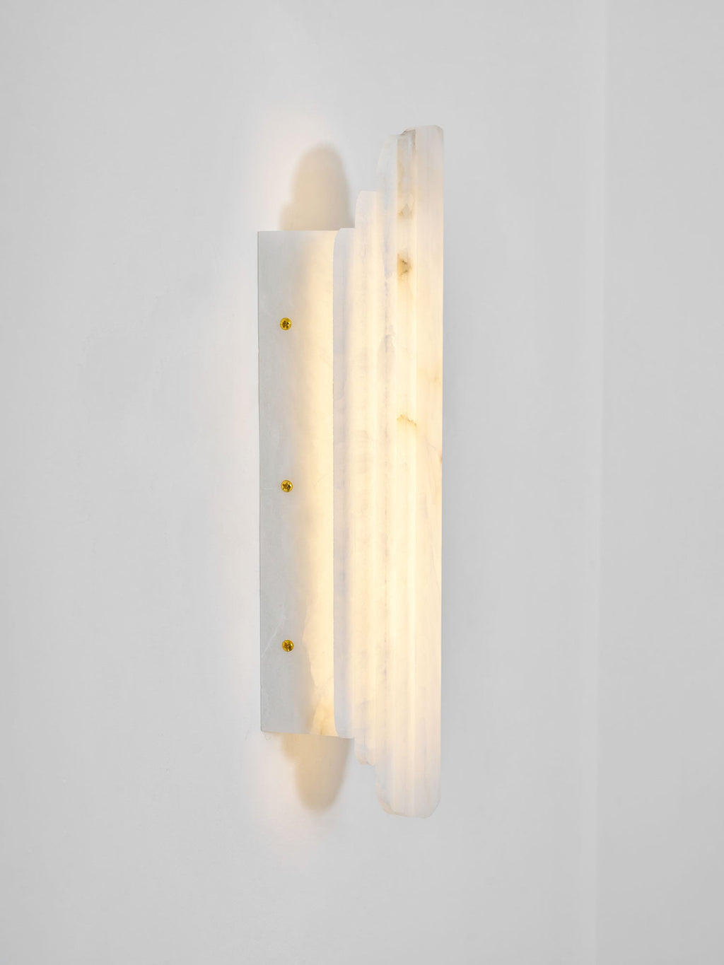 Side view of the lit Clérachic alabaster sconce, showing its stepped panels and a slim back plate with three brass-coloured screws.