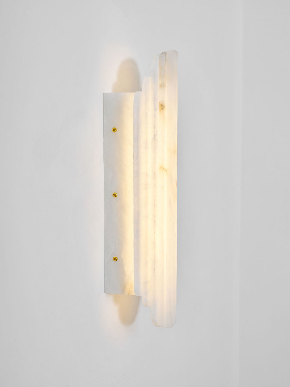 Side view of the lit Clérachic alabaster sconce, showing its stepped panels and a slim back plate with three brass-coloured screws.