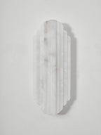 Front view of the unlit Clérachic alabaster sconce on a pale grey wall, showing the white, lightly veined ribs of the stone.