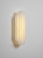 Three-quarter view of the lit Clérachic alabaster sconce on a pale wall, its ribbed shade casting a warm halo behind it.